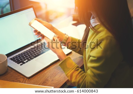Photo of Cropped image of young female skilled blogger holding modern smartphone downloading multimedia files from laptop computer with mock up monitor connected to free wireless internet in coworking space