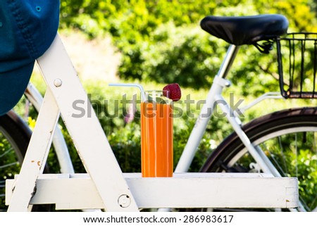 Glass of orange juice in the garden located on chair in shabby chic style. Blurred retro bike in the background surrounded by nature. Vacation and holiday atmosphere.