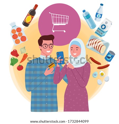 a Muslim couple do a groceries online shopping together on a mobile phone. The man is holding a card while the woman is holding a phone.