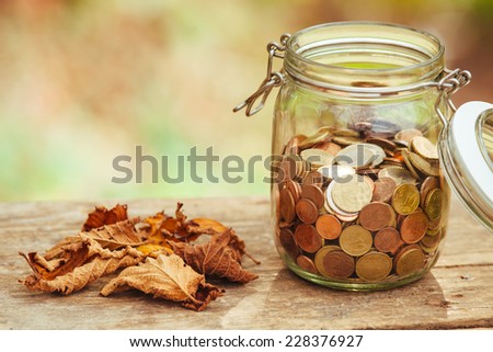 Money in a jar on a table next to some fallen leaves.