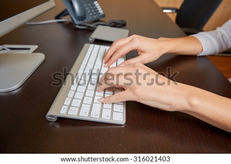 Woman typing on a computer keyboard on a wooden desk in front of a screen in the office.