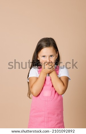 Portrait photo of female kid closing mouth with hands not talk with others with shining eyes and sullen look looking at camera wearing bright, pink jumpsuit and white t-shirt on beige background. Foto stock © 