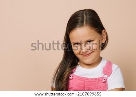 Close up portraitf female kid with shining bright eyes, humble smile and sullen look looking at camera wearing bright pink jumpsuit and white t-shirt on beige background. Foto stock © 