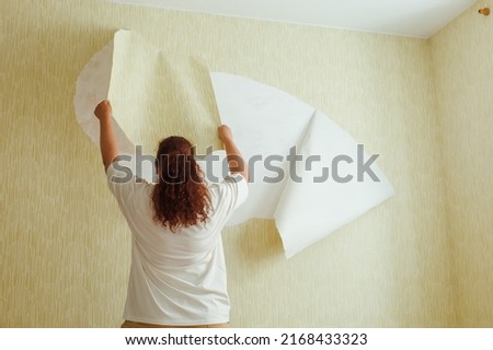 Back view of buxom woman with long curly red hair hair wearing white T-shirt, beige trousers, standing on chair, tearing off yellow wallpaper from wall. Home redecoration, interior change, renovation. Imagine de stoc © 