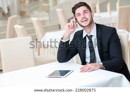 Good news on the phone. Young and motivated businessman is sitting at the table with tablet and mobile phone. Man is in a good mood and smiling, while doing his job