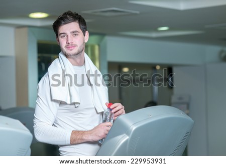 Portrait of young and handsome man in a gym. Man is standing with bottle of water in fitness gym and looking forward confidently
