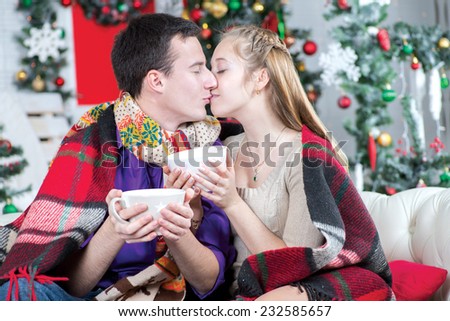 Christmas love. Young and beautiful pair is sitting on the sofa in festive New Year living room with cups of cacao and kissing each other before New Year