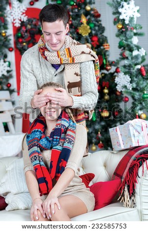 Ready to take Christmas presents. Loving couple is in the festive Christmas living room. Young and pretty girl is sitting on the sofa with closed eyes, while her husband is behind her