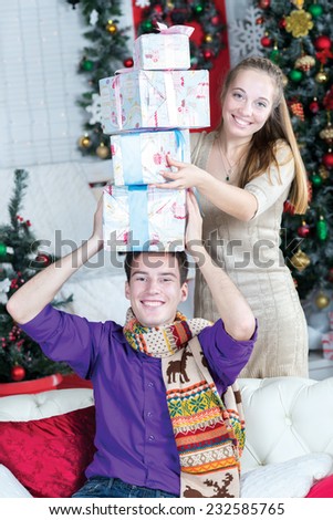 Young and beautiful pair is having fun with New Year presents in the living room. Husband is sitting on the sofa with presents on his head, while his wife is standing behind him and smiling