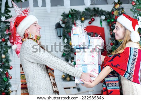 Merry Christmas and Happy New Year. Young and beautiful pair are standing in festive living room with presents and smiling to each other. Both are wearing New Year Santa hats