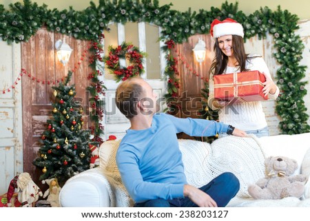 Christmas surprise. Couple in love is in festive Christmas decorated living room. Girl with smile is ready to give her husband a Christmas gift, while he is ready for New Year present.