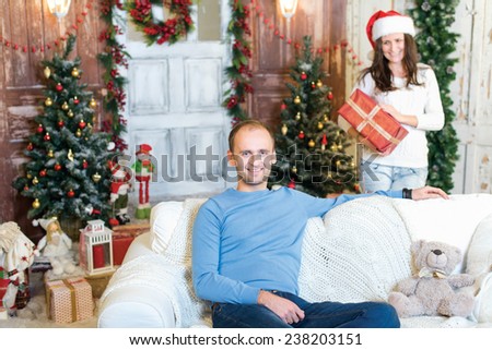 Christmas surprise. Couple in love is in festive Christmas decorated living room. Girl with smile is ready to give her husband a Christmas gift, while he is ready for New Year present