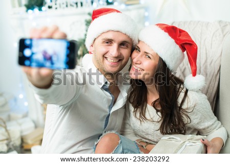 Christmas selfie. Couple in love is in festive Christmas decorated living room is making selfie on mobile phone just before happy New Year time. Both are wearing Santa Claus hats