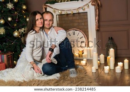 Merry Christmas and Happy New Year. Couple in love is sitting in festive Christmas decorated living room. Both are looking directly in the camera. Christmas background with candles and New Year tree
