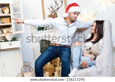 Merry Christmas and Happy New Year. Couple in love is in festive Christmas decorated living room. Girl is sitting in arm-chair, while her husband in Santa Claus hat is showing something