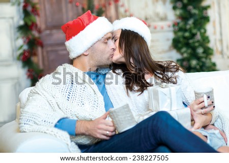 Merry Christmas and Happy New Year. Couple in love is sitting in festive Christmas decorated living room. Both are drinking New Year cacao from pretty Christmas cups and kissing each other