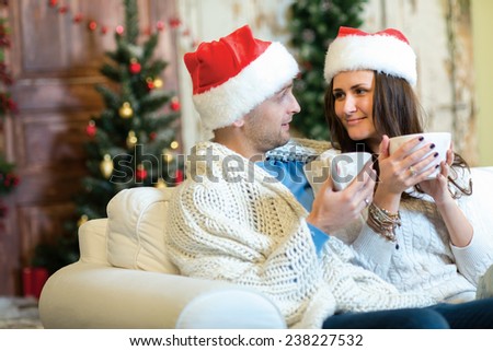 Merry Christmas and Happy New Year. Couple in love is sitting in festive Christmas decorated living room. Both are drinking New Year cacao from pretty Christmas cups and look at each other