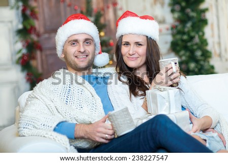 Merry Christmas and Happy New Year. Couple in love is sitting in festive Christmas decorated living room. Both are drinking New Year cacao from pretty Christmas cups and look in the camera