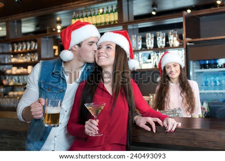 Love beer and Christmas. Loving couple is kissing in a bar or pub, wearing New Year Santa Claus hats. Girls bartender also is wearing Santa Christmas hat and stays on a background