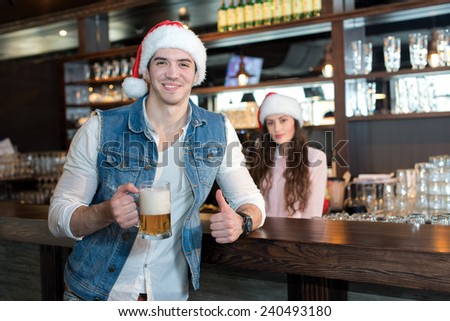 New Year with beer! Handsome guy is standing with beer in a bar or pub wearing Santa Claus New Year hat and smiling. Drinking beer before Christmas.