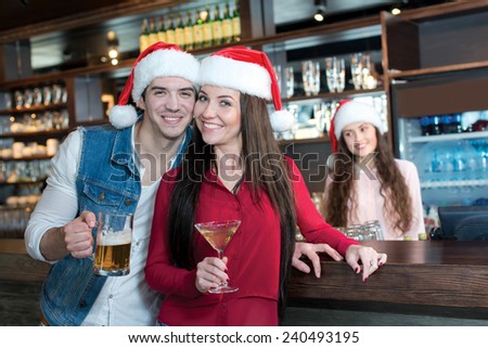 Love beer and Christmas. Loving couple is standing in a bar or pub, wearing New Year Santa Claus hats. Girls bartender also is wearing Santa Christmas hat and stays on a background