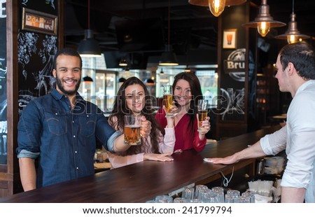 Perfect beer party. Portrait of young and handsome guy. He is standing at the bar counter and showing his beer. His female friends are standing with beer on the background and talking with each other