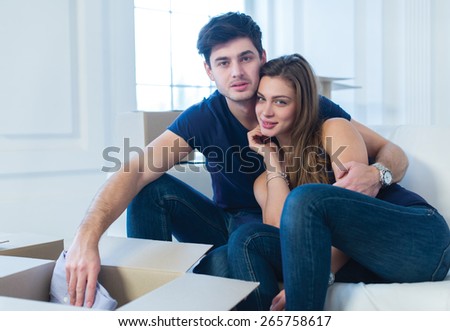Moving new flat with fun and excitement. Young and beautiful couple is moving to new apartment surrounded with plenty of cardboard boxes. Both are sitting on the sofa and unpacking boxes