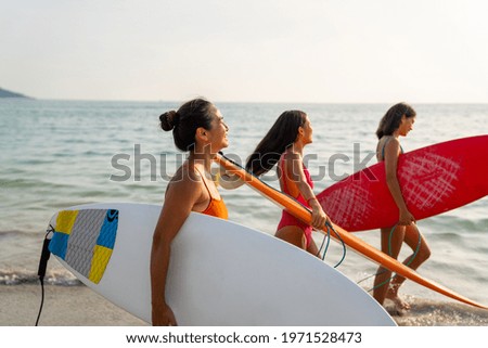 Group of Asian woman girl friends in swimwear holding surfboard and walking together on the beach at summer sunset. Female friendship enjoy outdoor activity lifestyle play extreme sports surfing