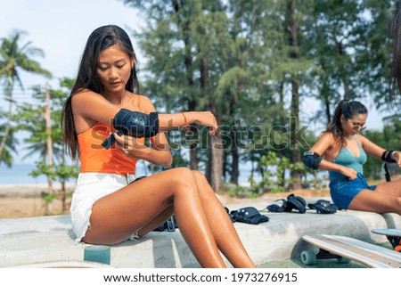 Asian woman with girl friends wear safety skateboarding arm pad before skating at skateboard park. Happy female friendship enjoy summer outdoor active lifestyle play extreme sport surf skate together