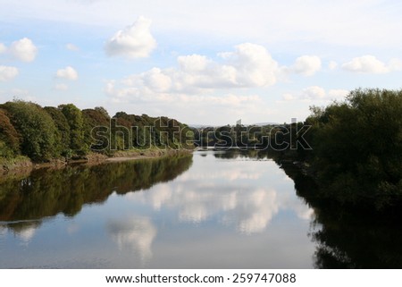 Calm waters of the tree lined River Ribble in Preston, Lancashire, reflecting clouds in the blue sky above.