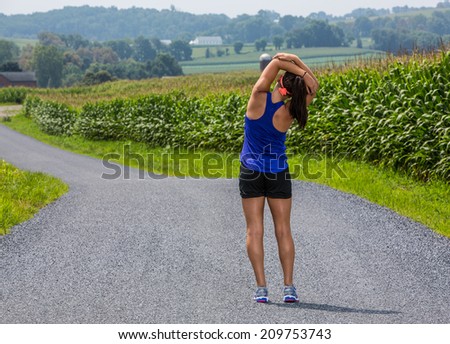 Young fit female athlete getting ready to run by stretching on a country road.