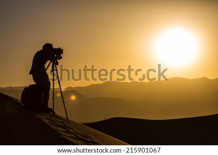 Photographer behind tripod on sand dune is silhouetted by setting sun in desert.