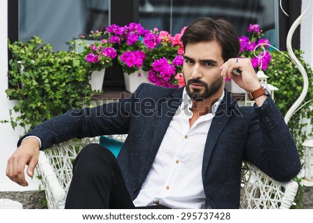 amazing man in a jacket and shirt sitting on a chair and looking forward, flowers background