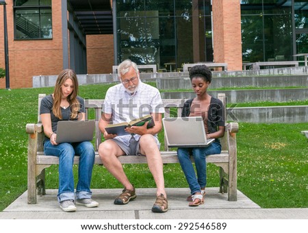 Old male student reading book with two mixed race young adult students with computers
