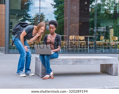 African American and Caucasian female college students sitting on bench interacting with laptop computer