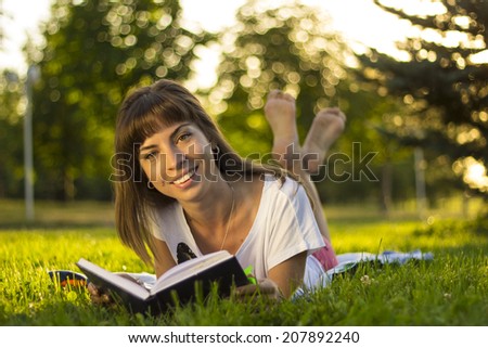 Beautiful smiling  young woman lying on grass and reading blue book, summer green park. Female student girl outside in park. Happy young university student of mixed European and Caucasian ethnicity.