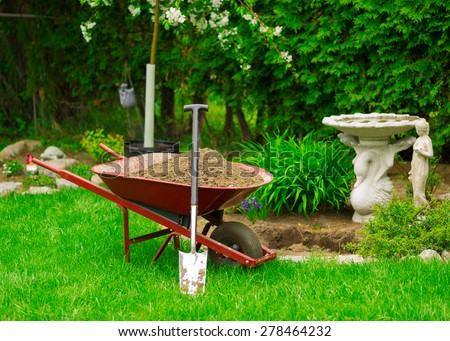 A red Wheelbarrow sits full of dirt in a lush, green garden as spring gardening work is underway.