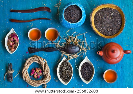 Clay teapot, three teacups, coil of rope, rosebuds, three wooden spoons, two handmade coconut bowls and three saucers in the form of petals with different sorts of tea on blue wooden background.