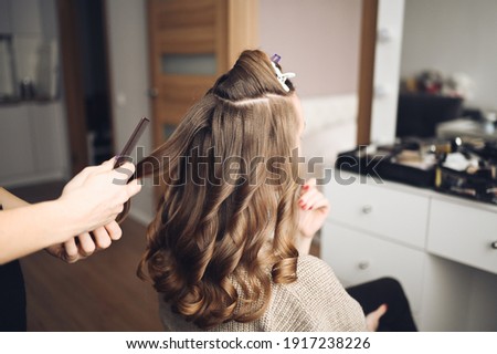 Hair stylist prepares woman makes curls hairstyle with curling iron. Long light brown natural hair