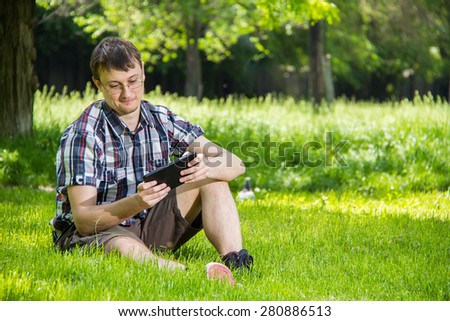 Young man in casual clothes uses digital tablet sitting on the grass in the park
