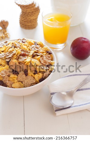 Delicious breakfast on a white wood table with a belly pitcher of milk, a bowl with cornflakes and raisins, a glass of orange juice, a pile of cookies, opened walnuts and a plum