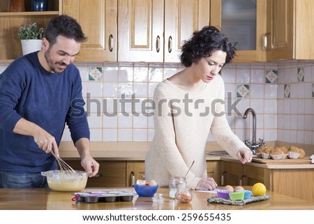 Happy couple cooking cupcakes in the kitchen. Man whisking dough and woman looking at recipe of homemade cupcakes