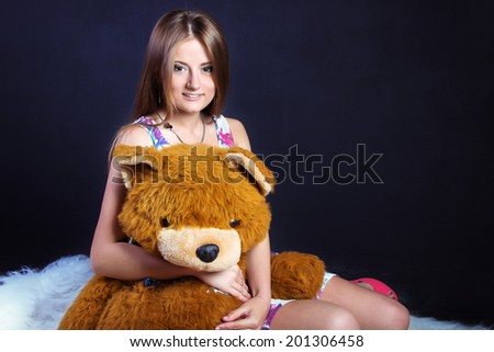 beautiful young girl with long hair with a smile in a dress with a large soft toy bear sitting on a white carpet in the Studio on a black background