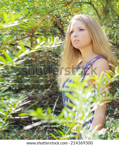 young girl with long blonde hair in a long Board is in among the bushes of sea-buckthorn in a Sunny bright day