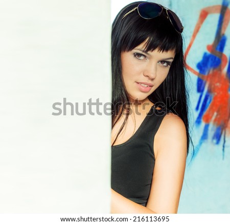 sexy beautiful girl with black hair and full lips, wearing sunglasses looks over the white wall