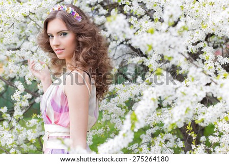 Pretty gentle young elegant beautiful girl with lush hair with a rim of brightly colored flowers in a garden near a flowering tree warm spring morning
