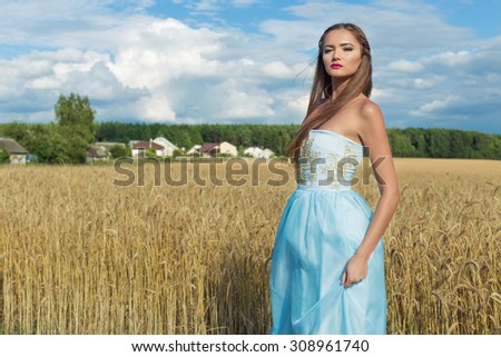 Young beautiful woman in a blue dress standing on a field of wheat against the blue sky