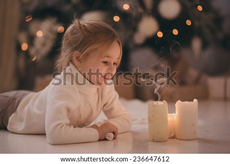 girl with blonde hair lying on the carpet blows out the candles. Christmas tree in the background. smiles
