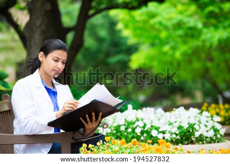 Closeup portrait, friendly, young serious confident female doctor, healthcare professional checking patient notes, bills isolated outside green trees, white flowers background. Health care reform.