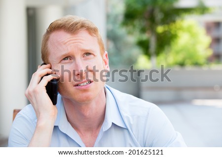 Closeup portrait, worried young man in blue shirt talking on phone to someone, looking up, isolated outdoors background